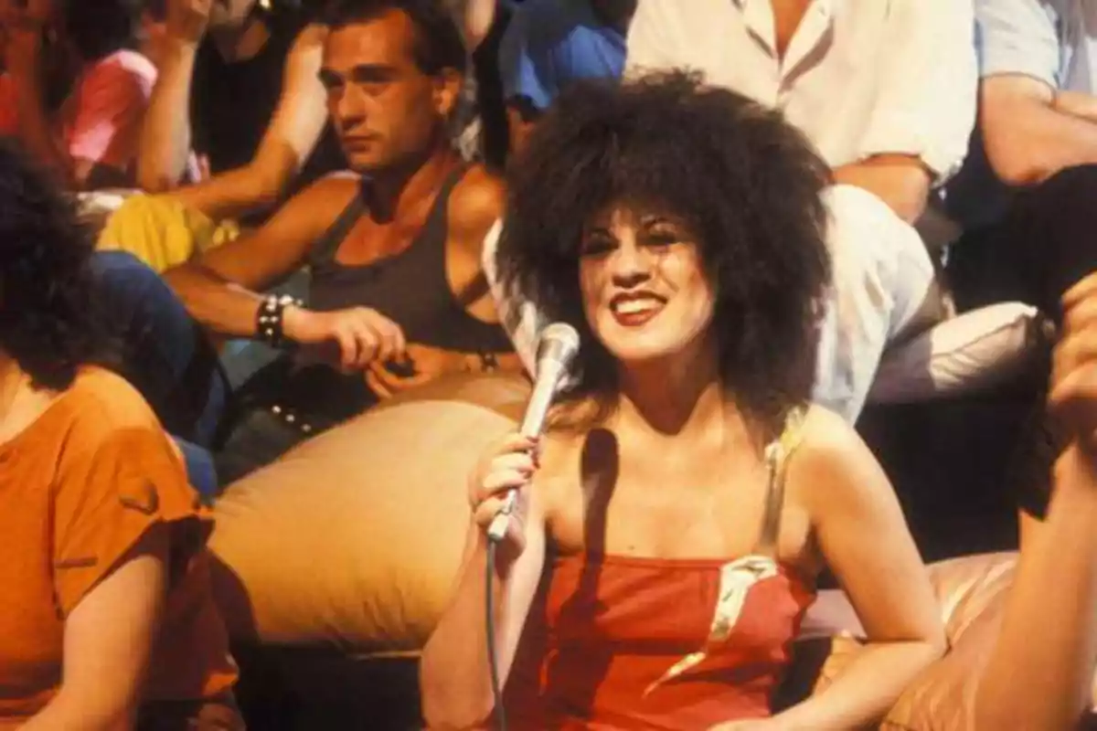 Una mujer con cabello rizado y vestido rojo sostiene un micrófono mientras sonríe, rodeada de personas sentadas en un ambiente informal.