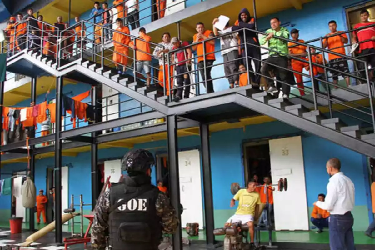 Un grupo de personas vestidas con uniformes naranjas se encuentran en un área de una prisión, distribuidos en diferentes niveles y escaleras, mientras un guardia con chaleco que dice "GOE" y otra persona observan la escena.
