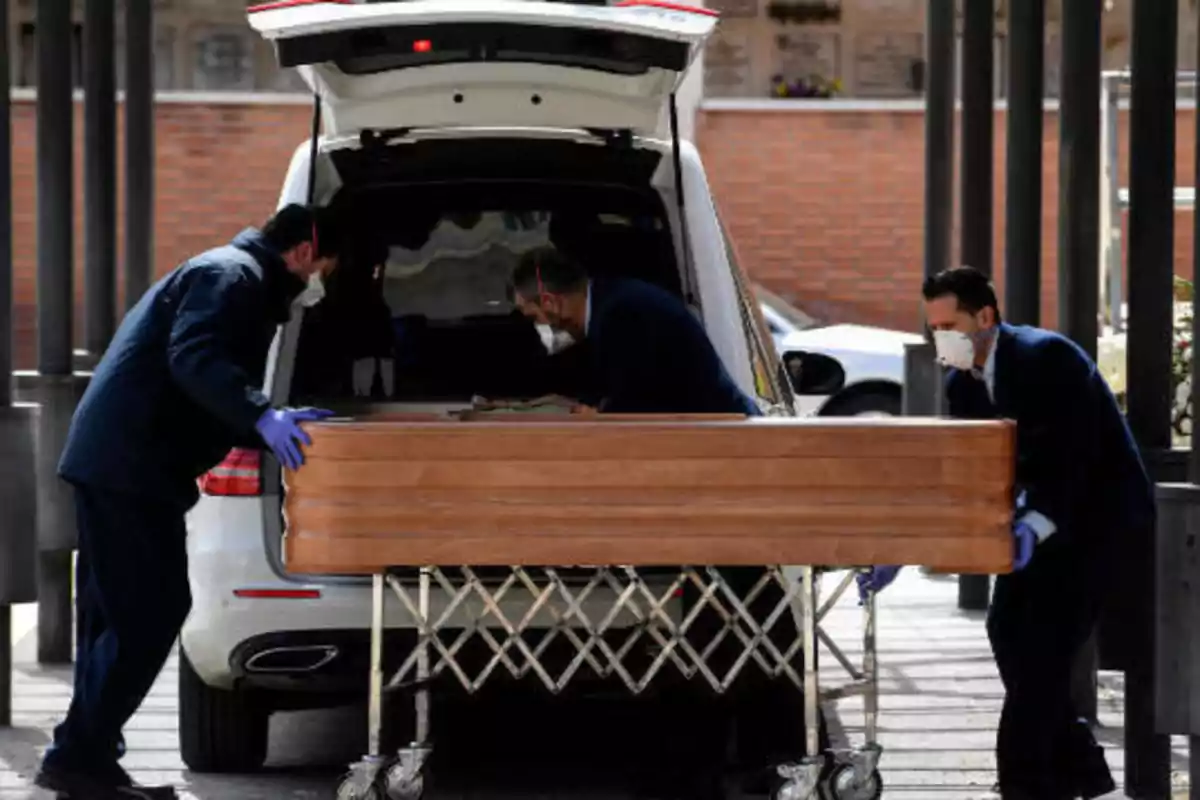 Tres personas con mascarillas y guantes colocan un ataúd en la parte trasera de un coche fúnebre.
