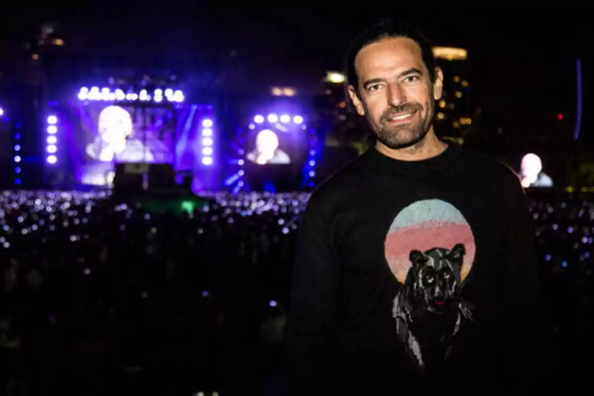 Hombre con suéter negro con diseño de pantera frente a un escenario iluminado en un concierto nocturno.
