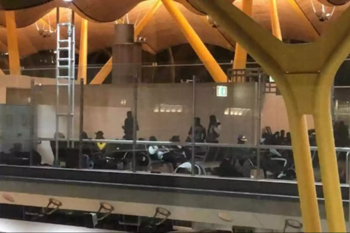 Personas esperando en una sala de un aeropuerto con estructura moderna y columnas amarillas.