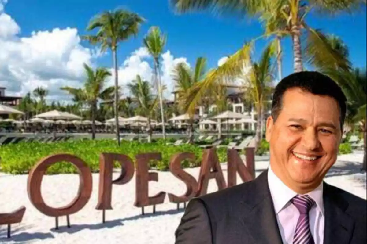Un hombre sonriente con traje y corbata está frente a un letrero que dice "LOPESAN" en una playa con palmeras y cielo despejado.