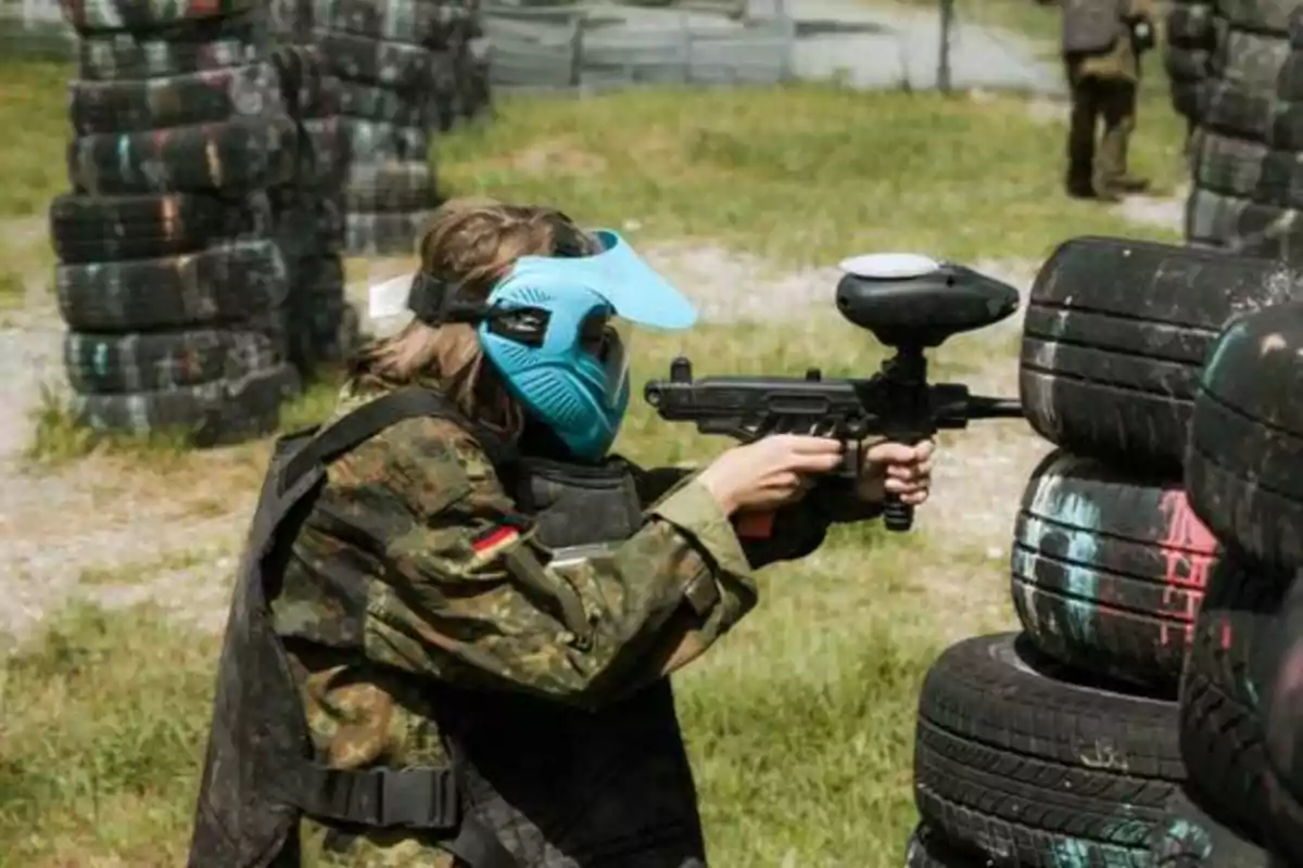 Persona con máscara azul y uniforme militar jugando paintball, apuntando con su marcador detrás de una pila de neumáticos.