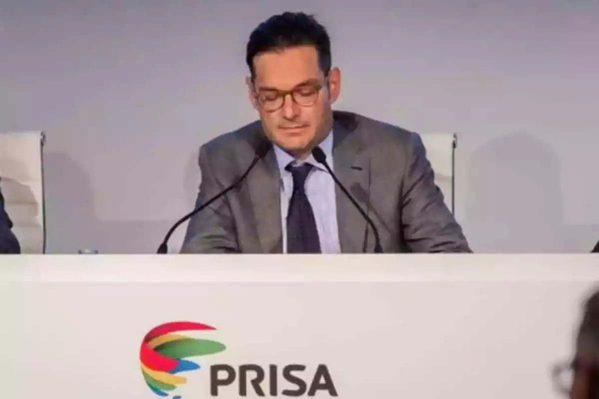 Hombre con gafas y traje sentado en una mesa de conferencia con el logo de PRISA al frente