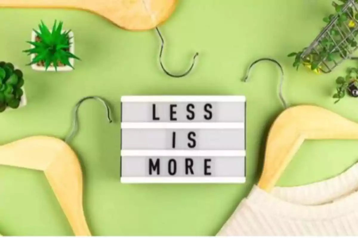 Un letrero con la frase "LESS IS MORE" rodeado de perchas de madera y plantas sobre un fondo verde.