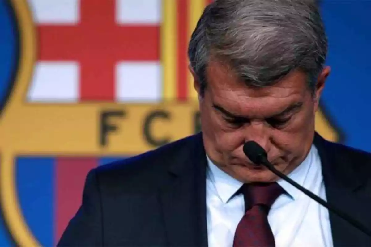 Un hombre con traje y corbata roja, con la cabeza inclinada hacia abajo, frente a un micrófono. Detrás de él, se ve el escudo del FC Barcelona.
