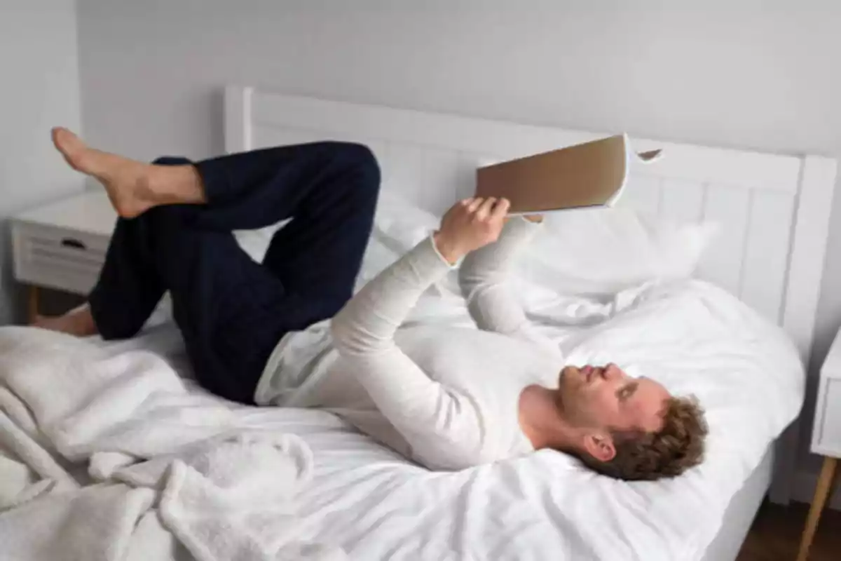Hombre acostado en una cama blanca leyendo un libro.