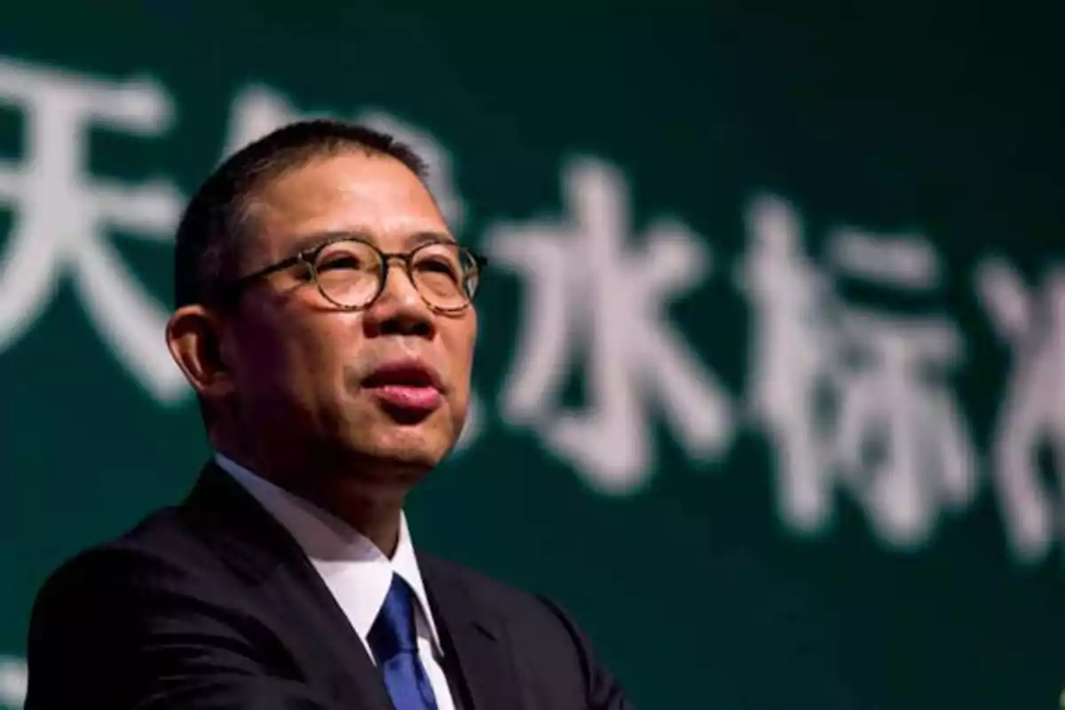 Hombre con gafas y traje oscuro hablando frente a un fondo verde con caracteres chinos.