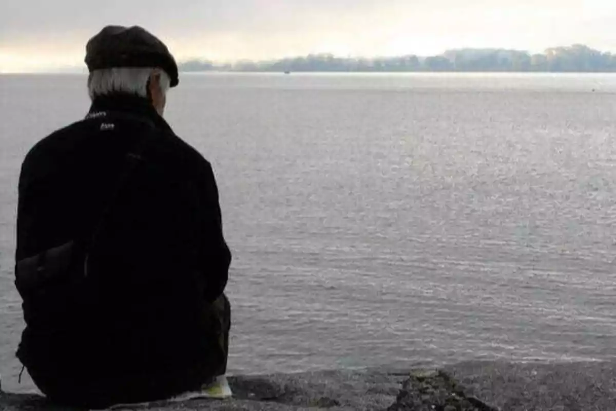 Un hombre mayor con gorra está sentado de espaldas, mirando hacia un lago tranquilo bajo un cielo nublado.