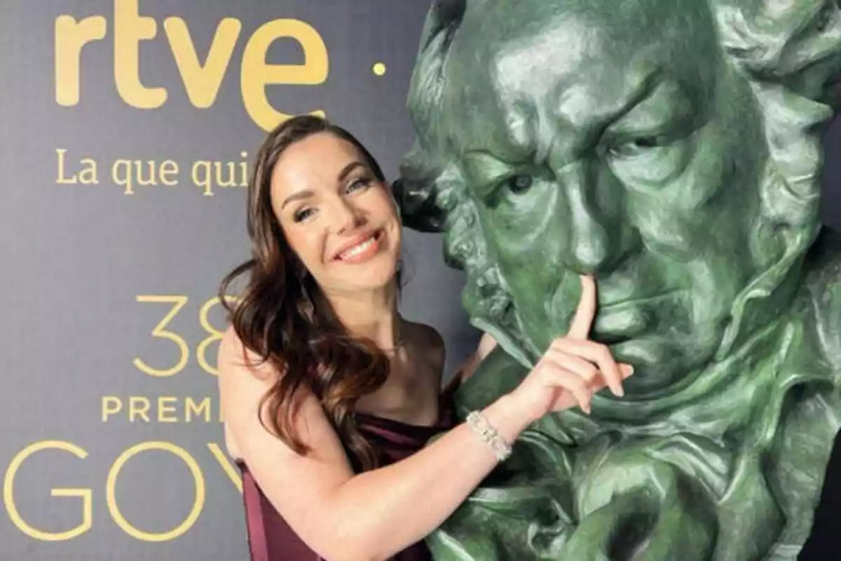 Una mujer sonriente posa junto a una estatua verde de un hombre, tocándole la nariz con su dedo índice. Al fondo se puede ver un cartel con el logo de RTVE y la frase "La que quieres", así como la mención a los "38 Premios Goya".