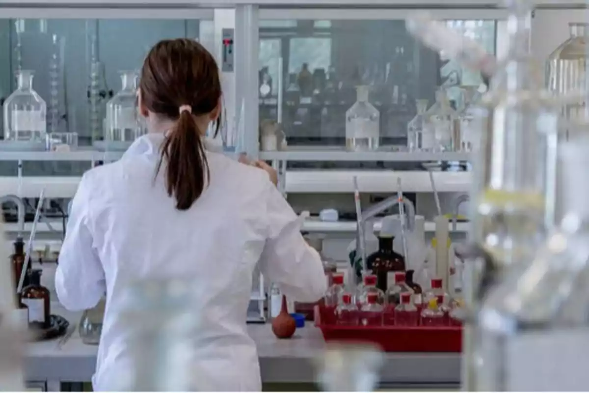 Persona con bata blanca trabajando en un laboratorio lleno de frascos y equipos científicos.