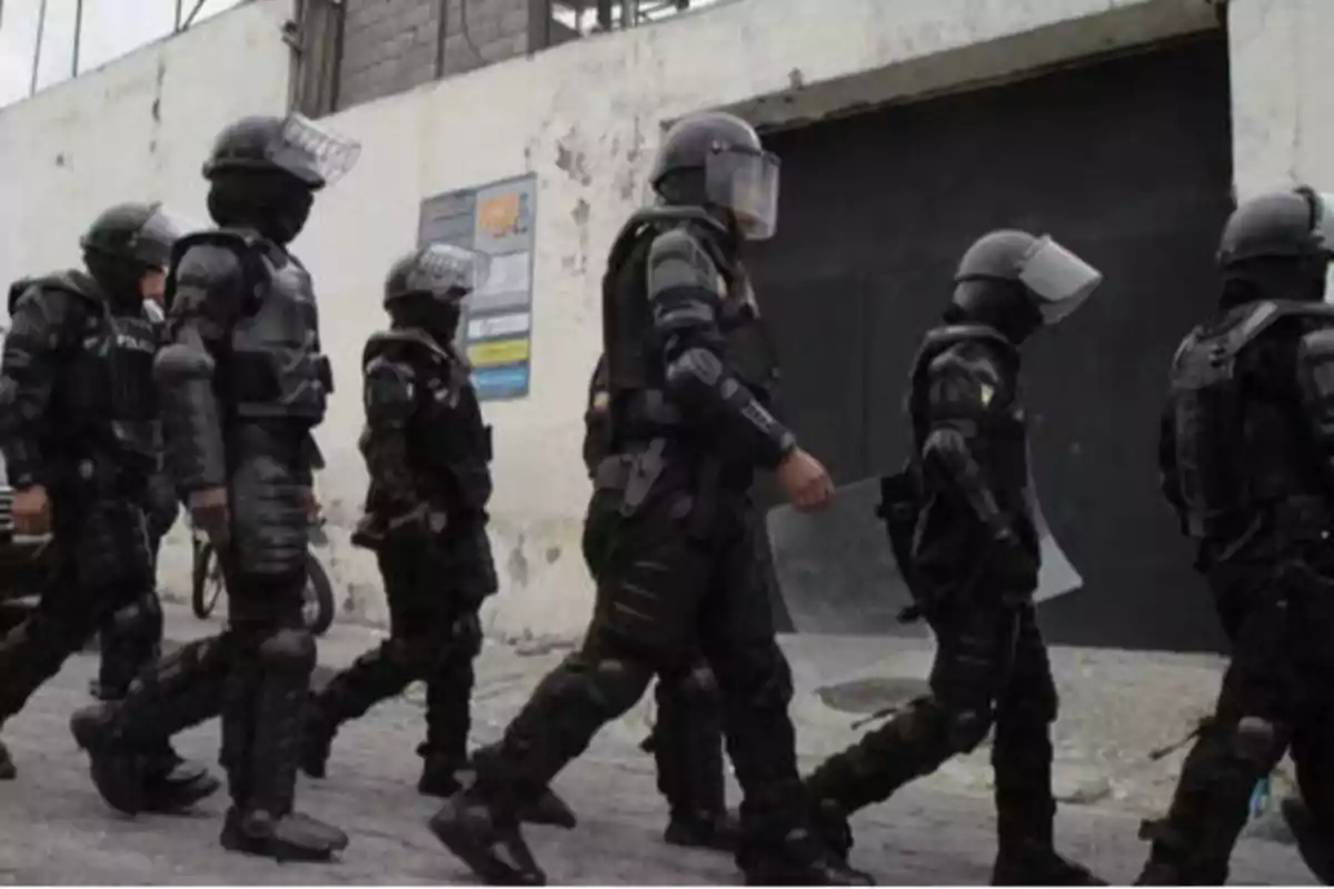 "Un grupo de policías antidisturbios con equipo de protección completo, incluyendo cascos y escudos, caminando en fila frente a un edificio con paredes blancas y desgastadas."