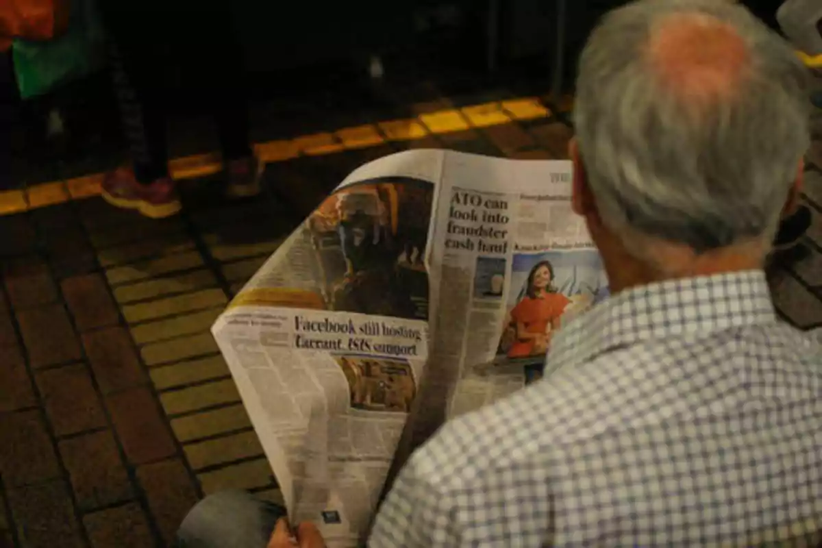 Un hombre de cabello canoso y camisa a cuadros lee un periódico en un lugar con suelo de baldosas.