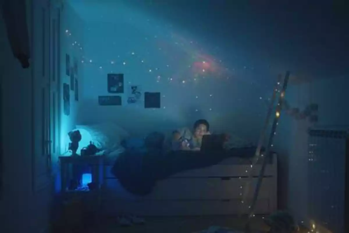 Una persona acostada en una cama en una habitación oscura iluminada por luces de colores y una computadora portátil.
