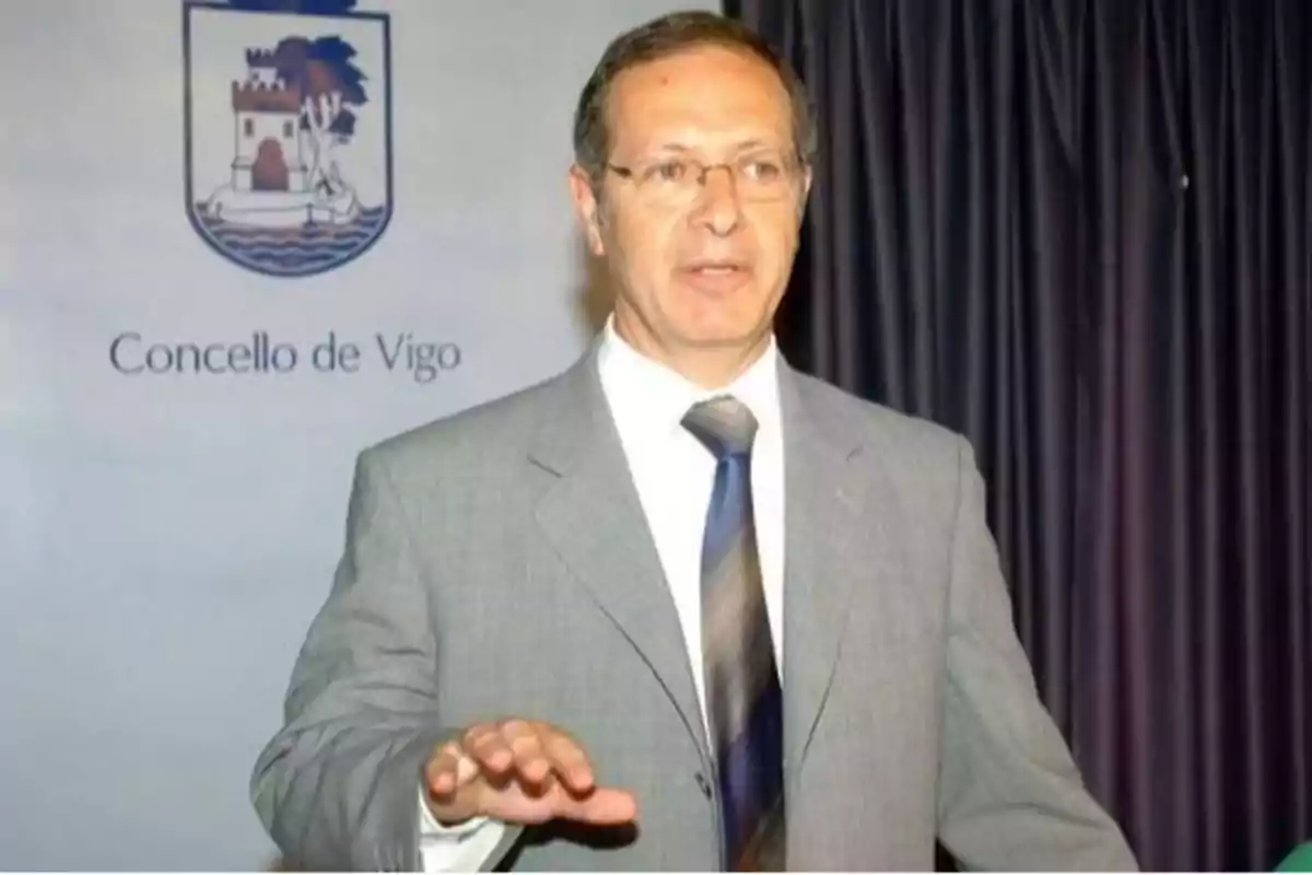 Hombre con traje gris y corbata azul hablando frente a un fondo con el escudo y el nombre del Concello de Vigo.