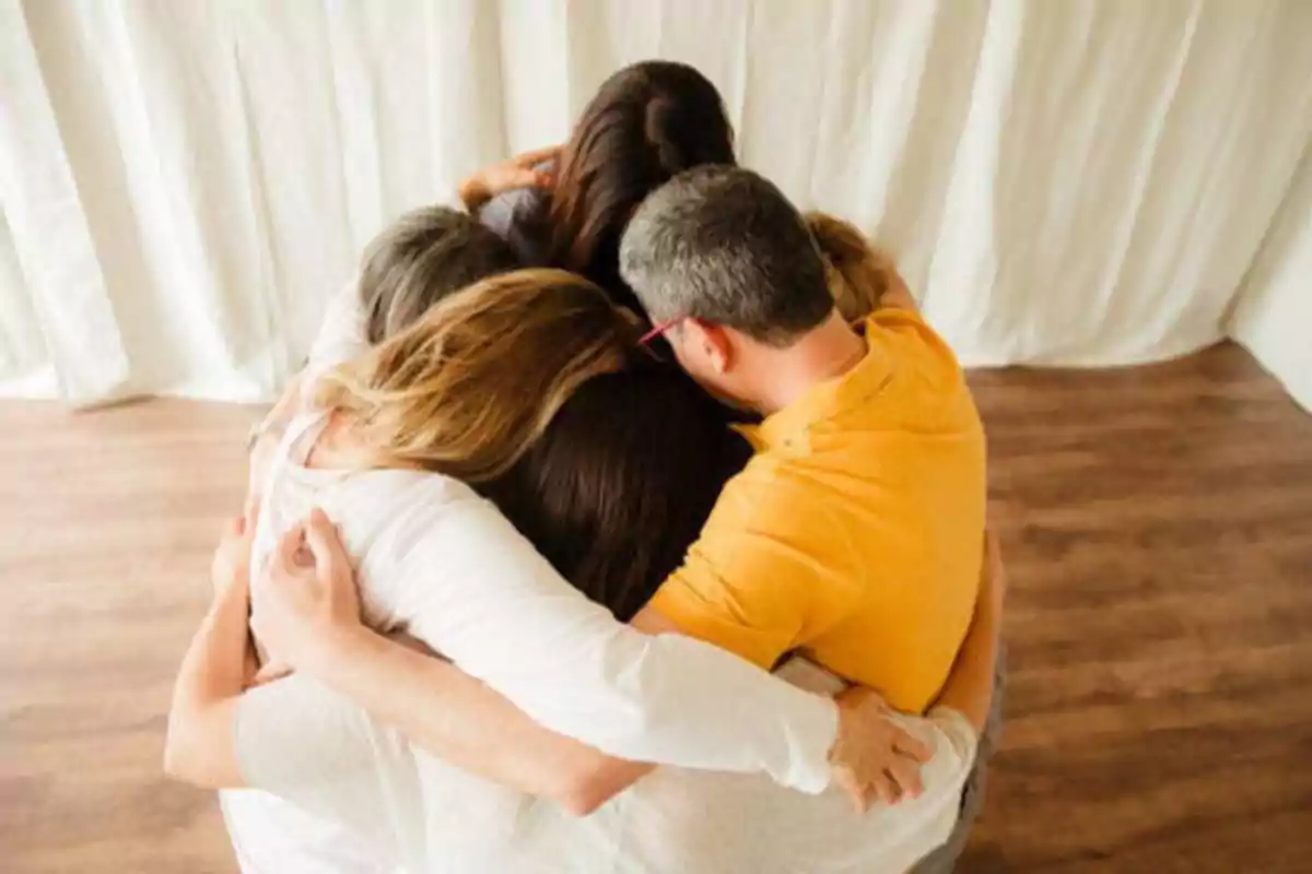 Un grupo de personas abrazándose en círculo sobre un suelo de madera con cortinas blancas de fondo.