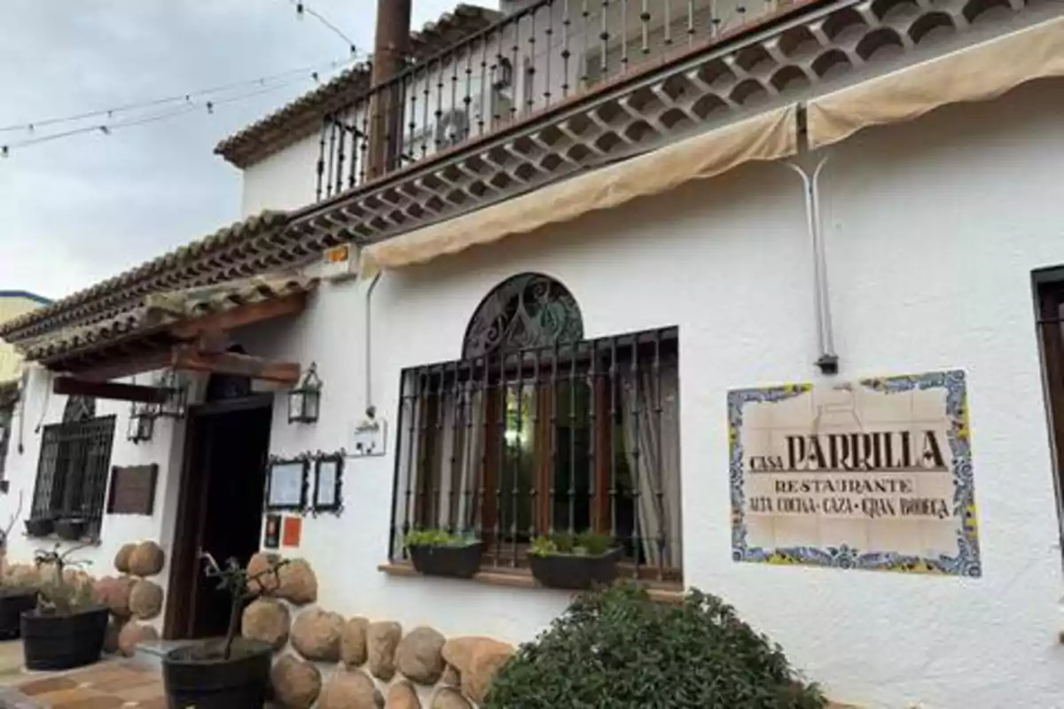 Fachada de un restaurante con paredes blancas, ventanas con rejas y un letrero que dice "Casa Parrilla Restaurante Alta Cocina Caza Gran Bodega".
