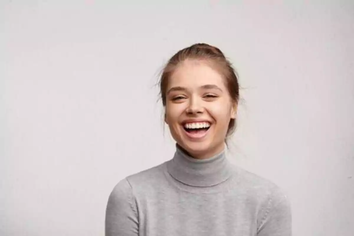 Persona joven sonriendo con un suéter gris de cuello alto y fondo claro.