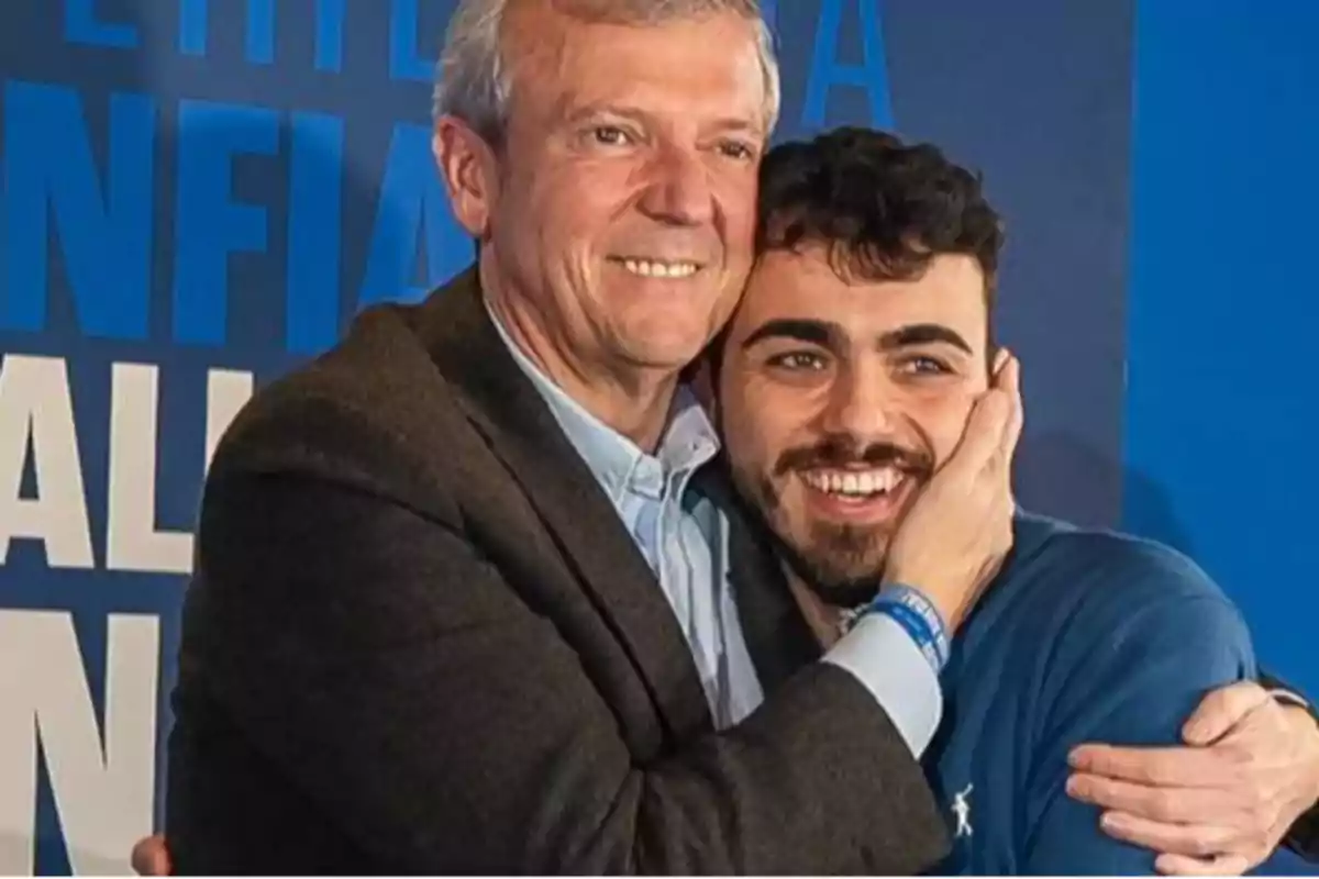 Dos hombres sonrientes se abrazan frente a un fondo azul con texto.
