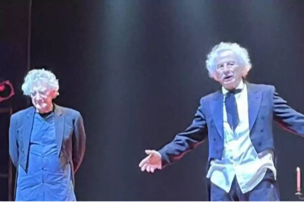 Dos hombres mayores con cabello canoso y rizado están en un escenario, uno de ellos lleva un traje y el otro una chaqueta casual.