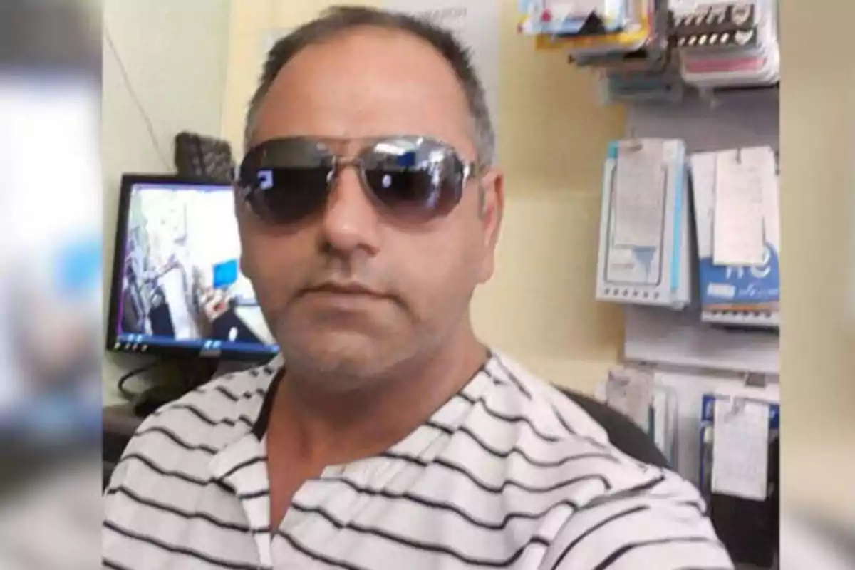 Hombre con gafas de sol y camiseta de rayas posando frente a una computadora en una oficina.