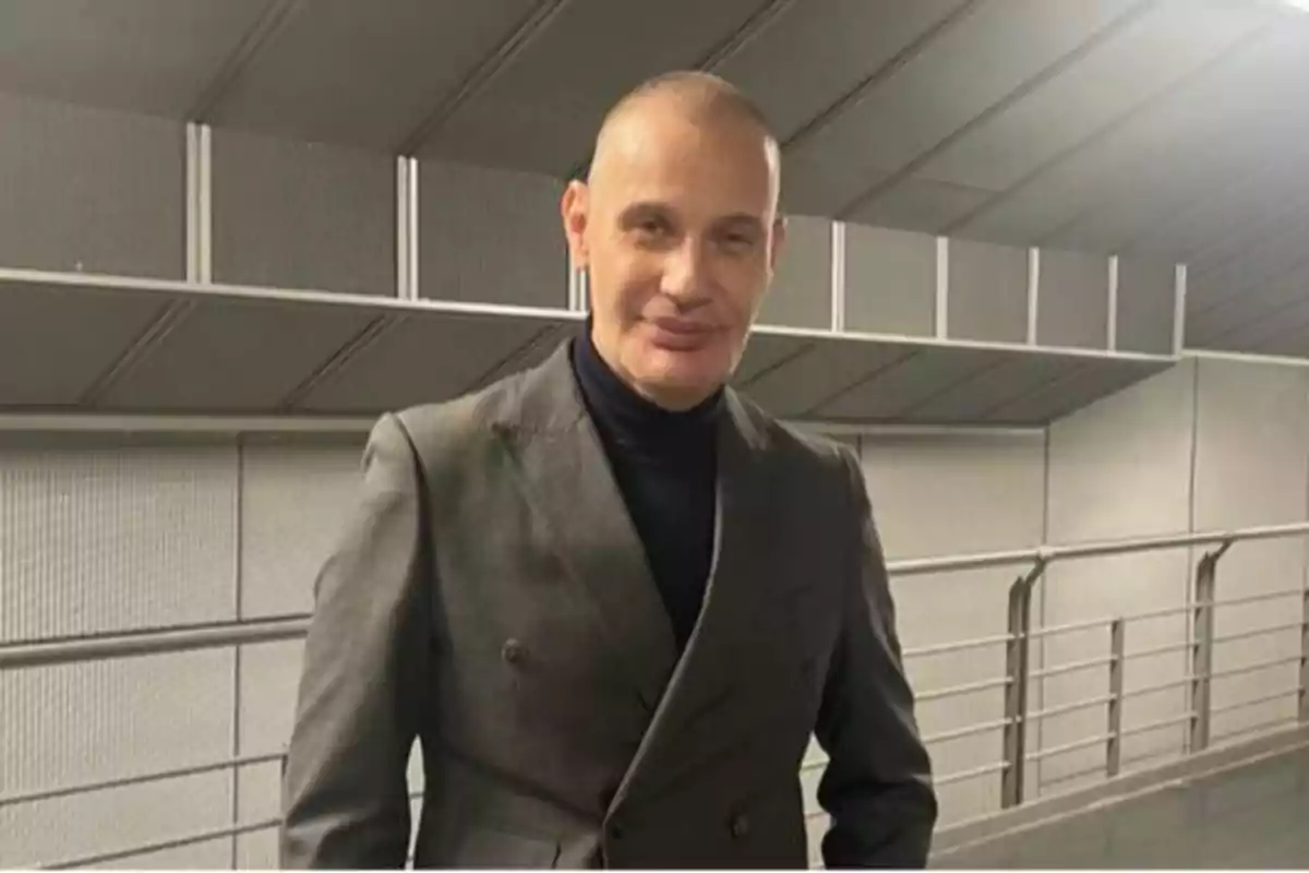 Un hombre calvo con traje gris y suéter negro posando en un entorno moderno con barandales y paredes grises.