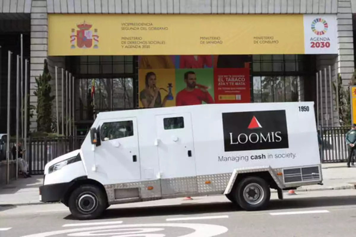 Un camión blindado de la empresa Loomis está estacionado frente a un edificio gubernamental con un cartel que dice "Vicepresidencia Segunda del Gobierno, Ministerio de Derechos Sociales y Agenda 2030, Ministerio de Sanidad, Ministerio de Consumo".