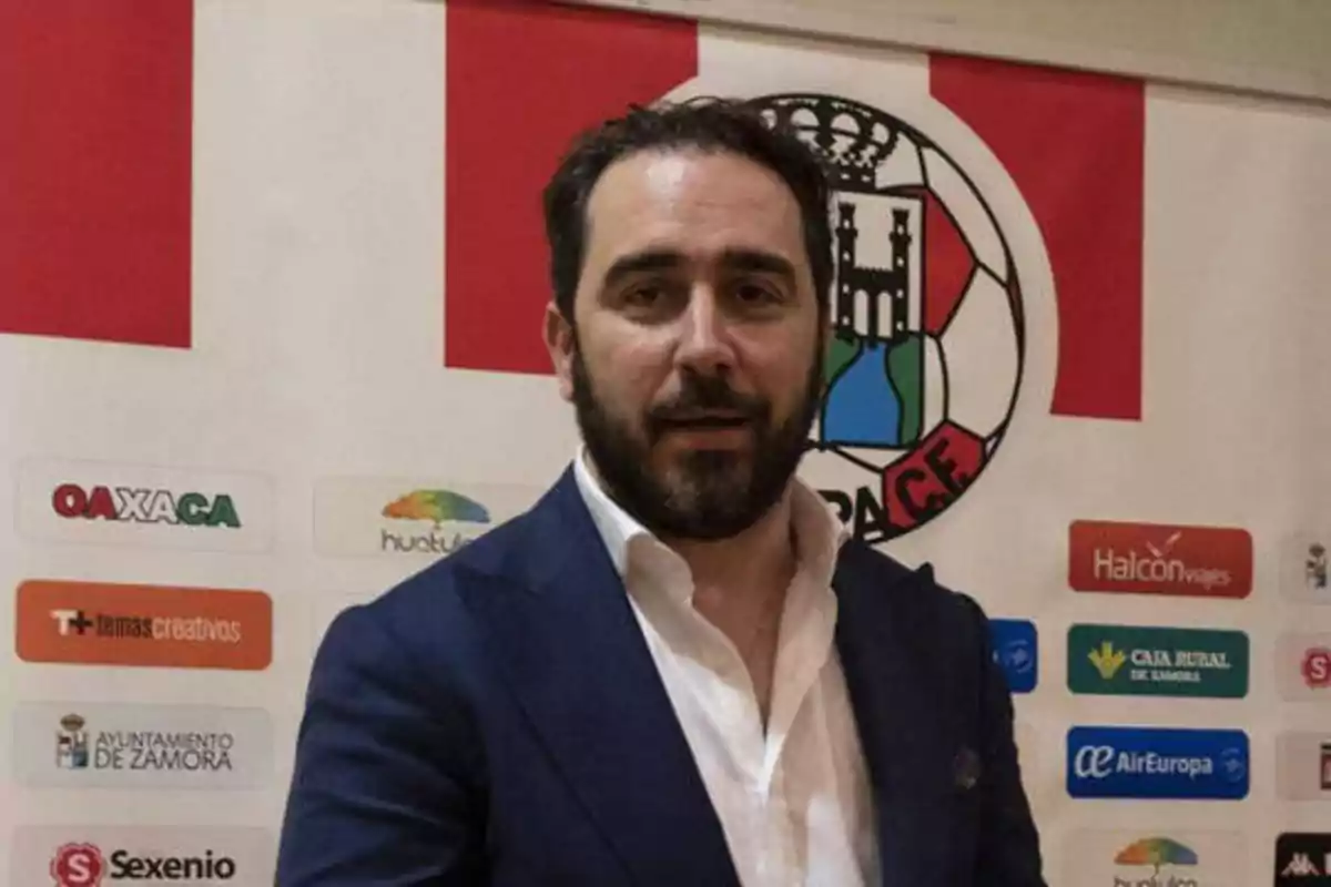 Un hombre con barba y traje oscuro está de pie frente a un fondo con logotipos de patrocinadores y un escudo de un equipo de fútbol.