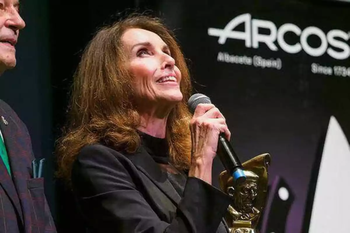 Una mujer sosteniendo un micrófono y un trofeo mientras sonríe en un evento.