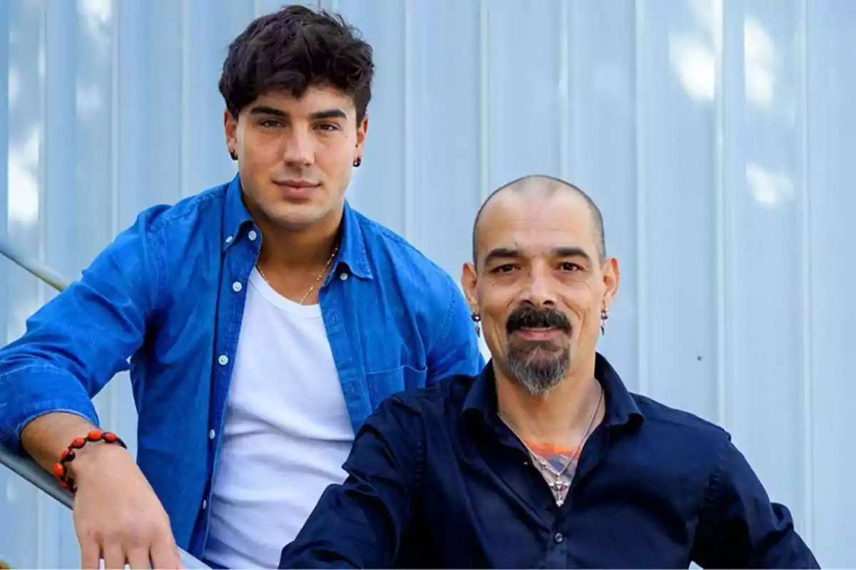 Dos hombres posando juntos, uno joven con camisa azul y el otro mayor con camisa negra.