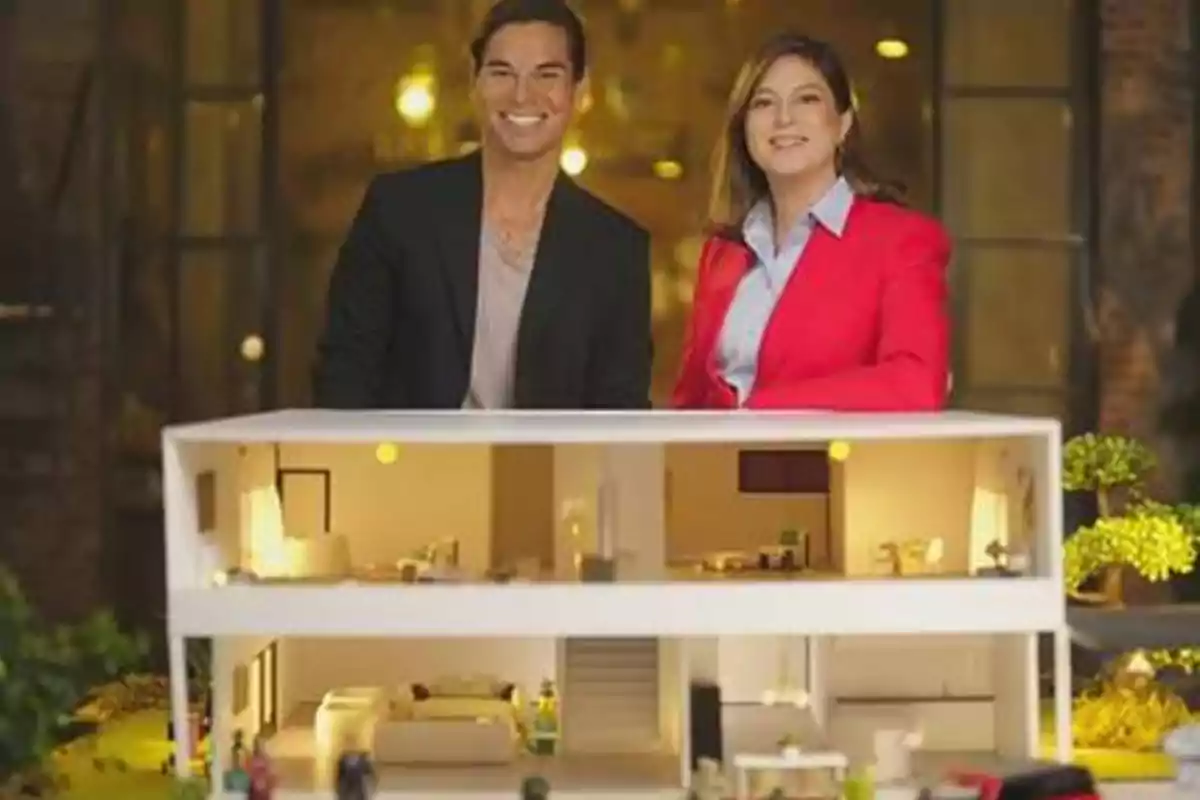 Dos personas sonrientes posan detrás de una maqueta de una casa moderna iluminada.