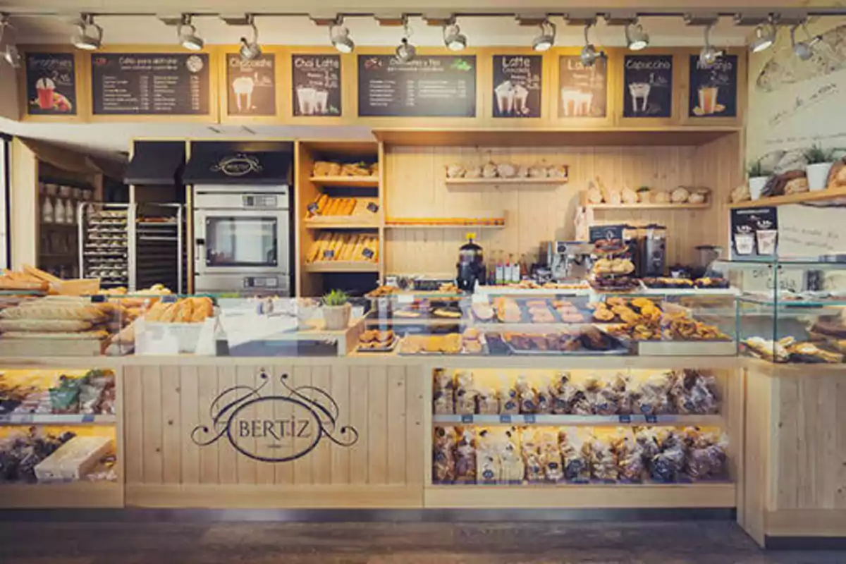 Una panadería moderna con un mostrador de madera, exhibiendo una variedad de panes y pasteles, con un menú de bebidas en la parte superior y estantes llenos de productos horneados.