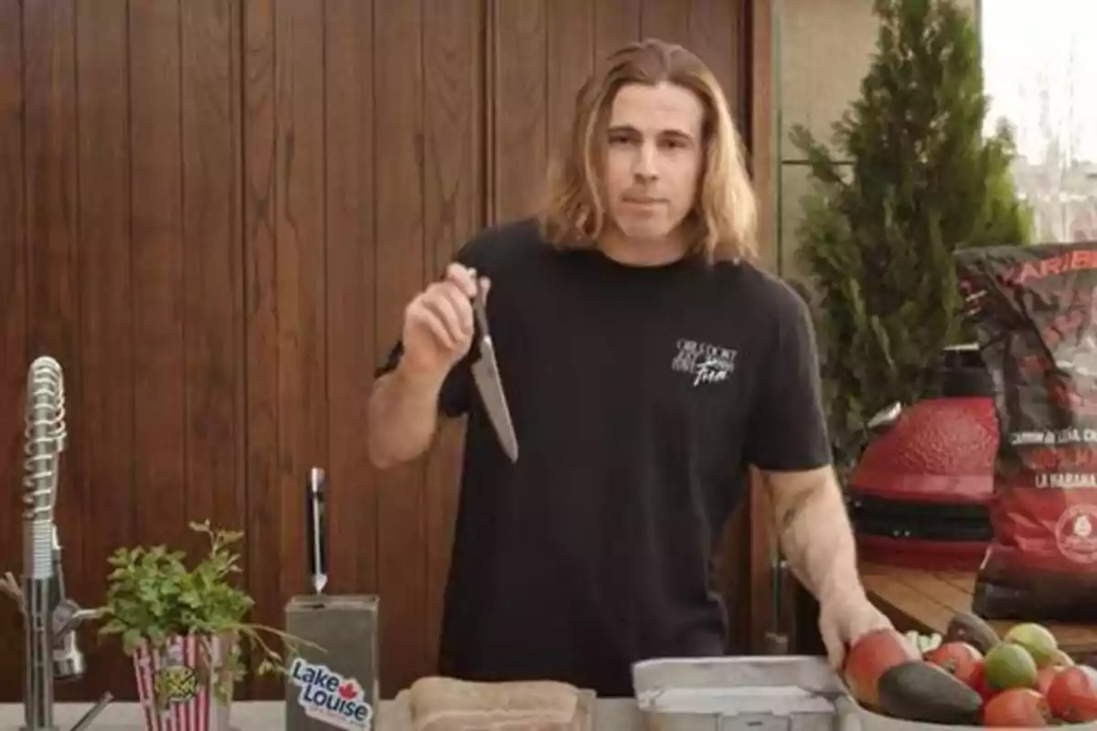Un hombre con cabello largo y una camiseta negra sostiene un cuchillo en una cocina al aire libre, con una tabla de cortar, un recipiente con aguacates y tomates, y una planta en una maceta decorativa.