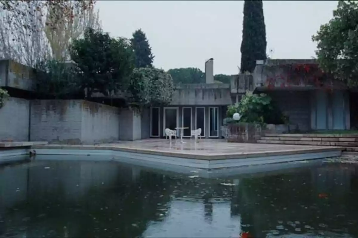 Una casa moderna con paredes de concreto, rodeada de vegetación, con una piscina en primer plano y un patio con sillas y una mesa.