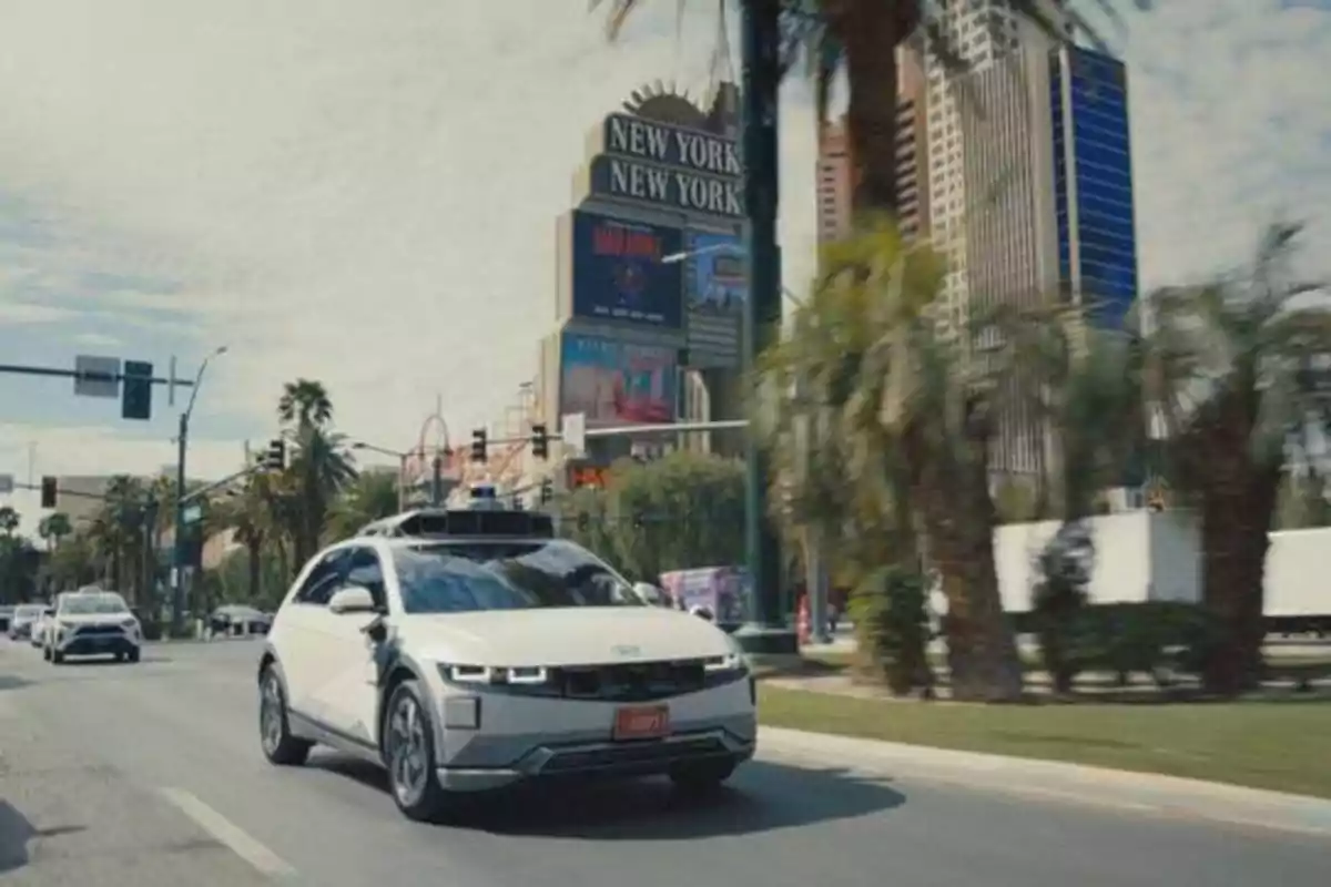 Un coche blanco circula por una calle rodeada de palmeras y edificios altos, con un cartel que dice "New York New York" en el fondo.