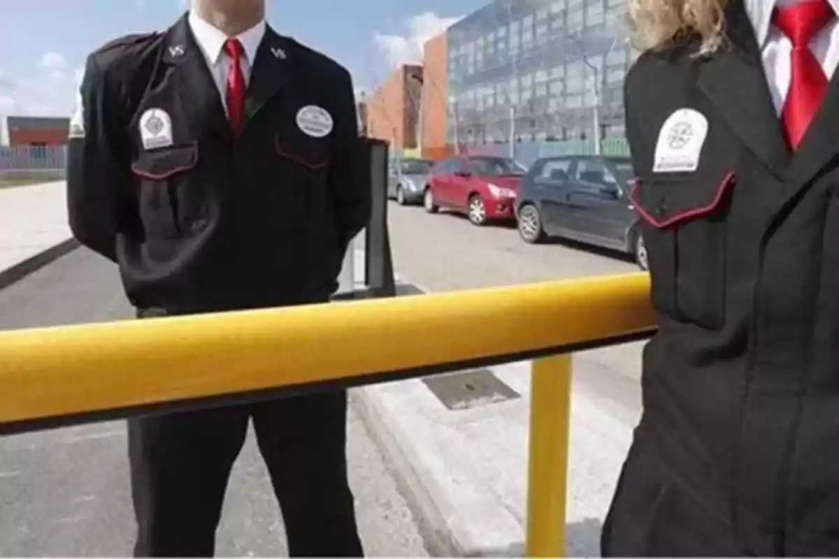 Dos guardias de seguridad con uniformes negros y corbatas rojas están de pie detrás de una barrera amarilla en un estacionamiento con varios autos y un edificio de fondo.