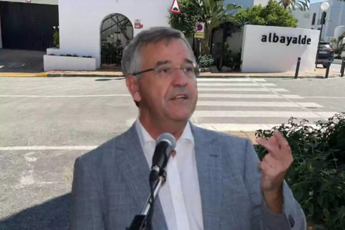 Hombre con traje gris hablando en un micrófono frente a un edificio blanco con el letrero "albayalde".