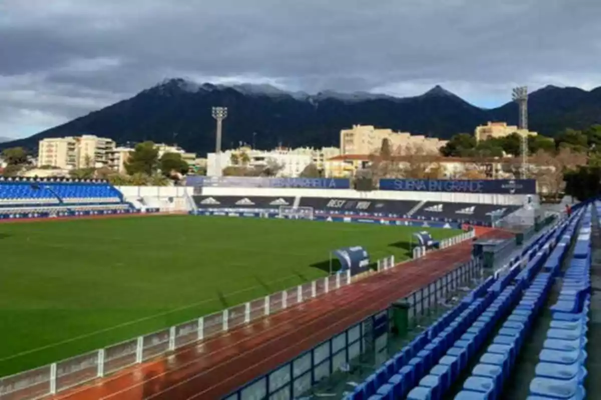 Estadio de fútbol con gradas azules y pista de atletismo, rodeado de edificios y montañas al fondo.