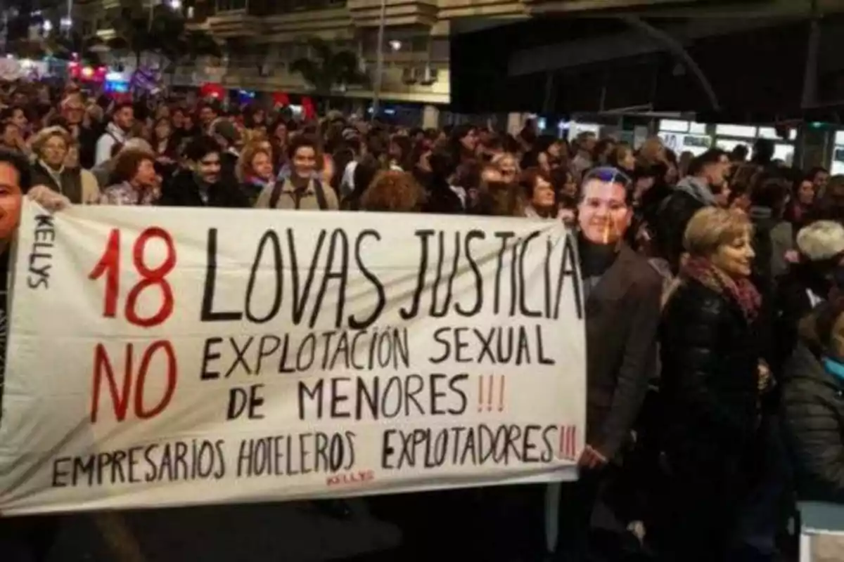 Una multitud de personas se manifiesta en la calle sosteniendo una pancarta que dice "18 LOVAS JUSTICIA NO EXPLOTACIÓN SEXUAL DE MENORES!!! EMPRESARIOS HOTELEROS EXPLOTADORES!!!"