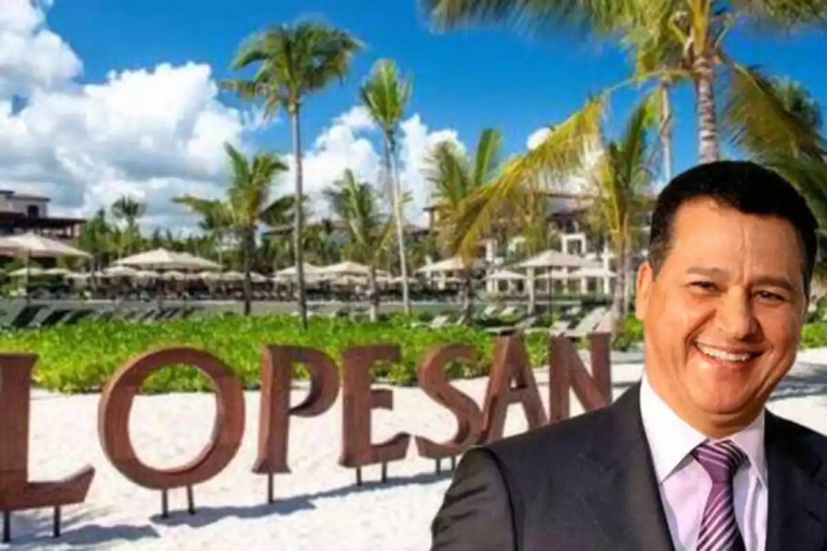 Hombre sonriendo frente a un letrero de "LOPESAN" en una playa con palmeras y cielo despejado.