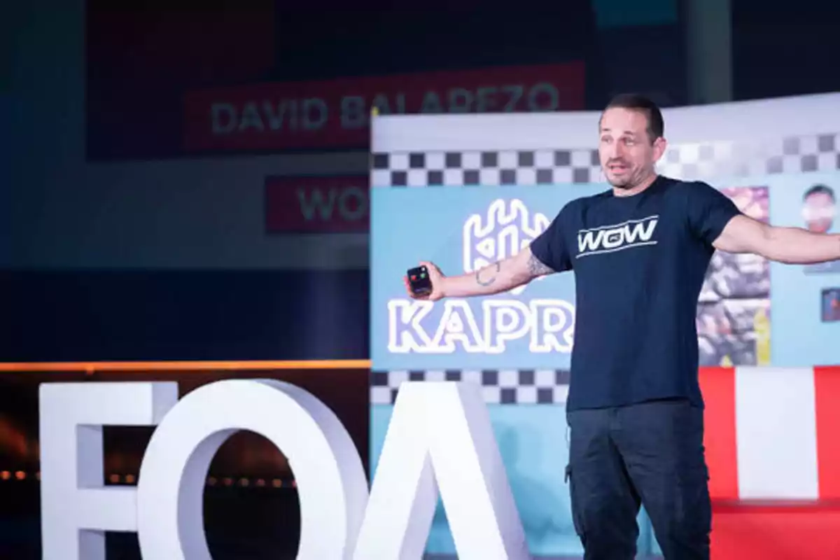 Un hombre con una camiseta negra y pantalones oscuros está de pie en un escenario con los brazos extendidos, detrás de él hay un cartel con el texto "KAPRO" y letras grandes que forman "FOA".