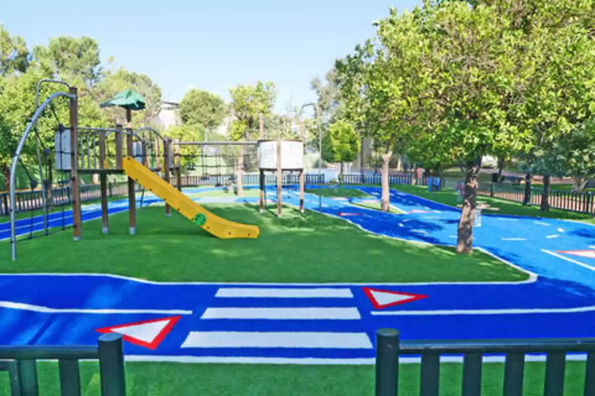 Un parque infantil con un tobogán amarillo, estructuras de juego de madera y un área de juegos con caminos azules y marcas viales blancas, rodeado de árboles y césped artificial.