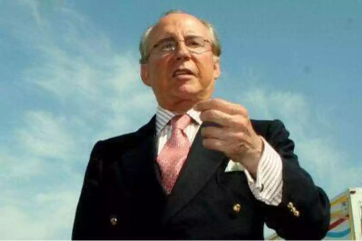Hombre de mediana edad con gafas y traje oscuro, corbata rosa y camisa blanca, gesticulando con la mano, con un cielo azul de fondo.