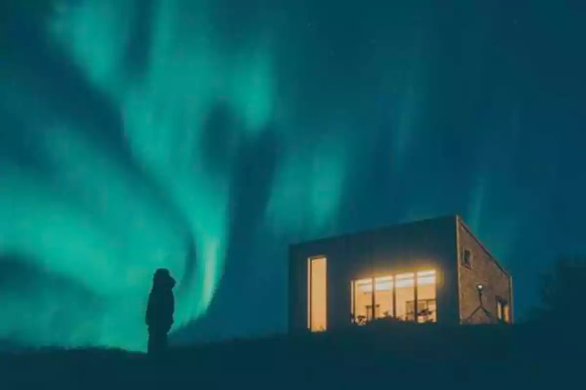 Una persona de pie en la oscuridad observa una casa iluminada con la aurora boreal en el cielo.
