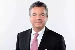 Hombre de mediana edad con traje negro, camisa blanca y corbata rosa, posando frente a un fondo blanco.