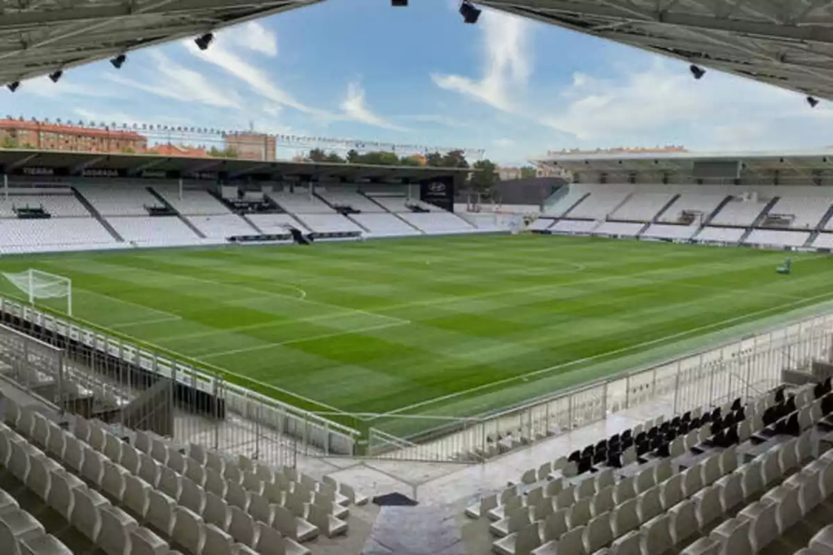 Estadio de fútbol vacío con gradas blancas y césped verde bajo un cielo parcialmente nublado.