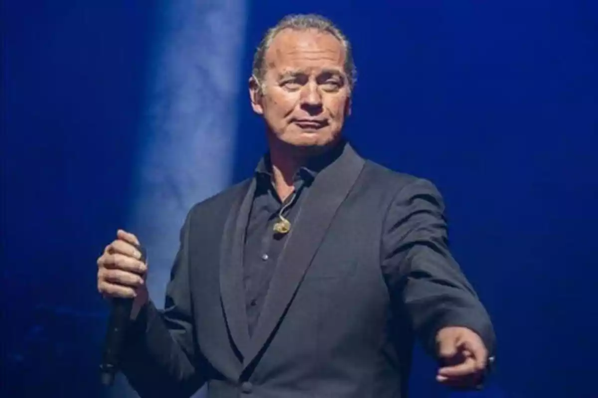 Hombre de pie en el escenario con un micrófono en la mano, vestido con un traje oscuro y fondo azul.