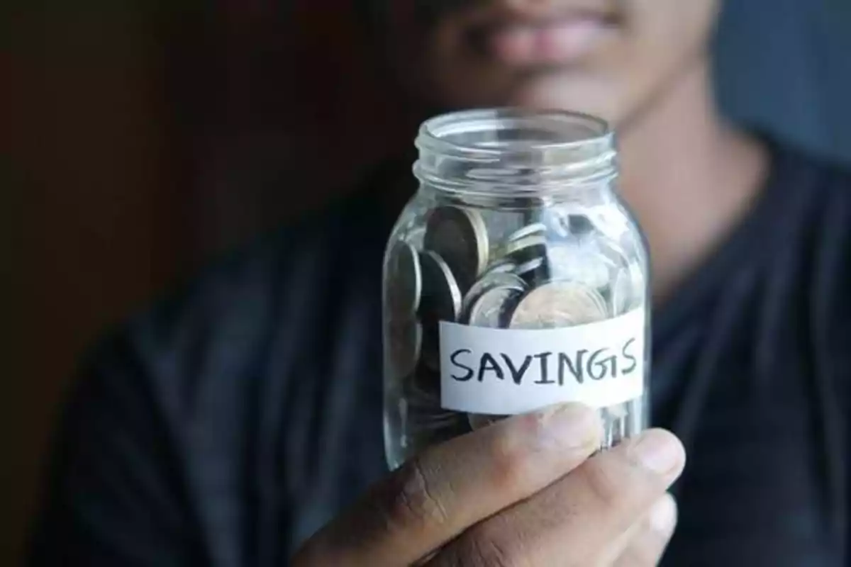 Persona sosteniendo un frasco de vidrio lleno de monedas con una etiqueta que dice "SAVINGS"