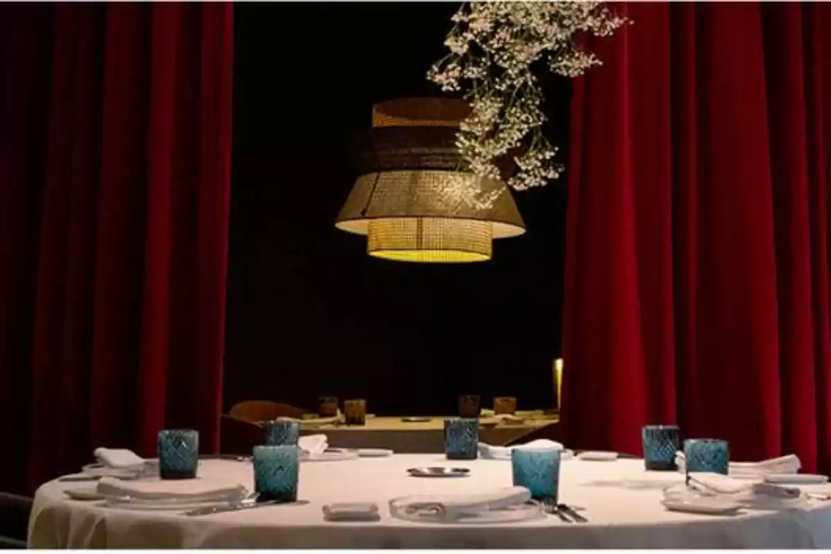 Mesa de comedor elegantemente puesta con copas azules y servilletas blancas, rodeada de cortinas rojas y una lámpara colgante decorativa.
