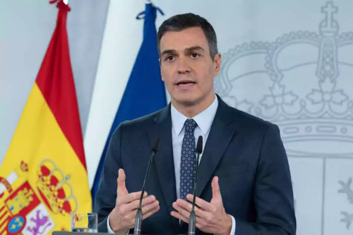 Hombre en traje dando un discurso con banderas de España y la Unión Europea de fondo.